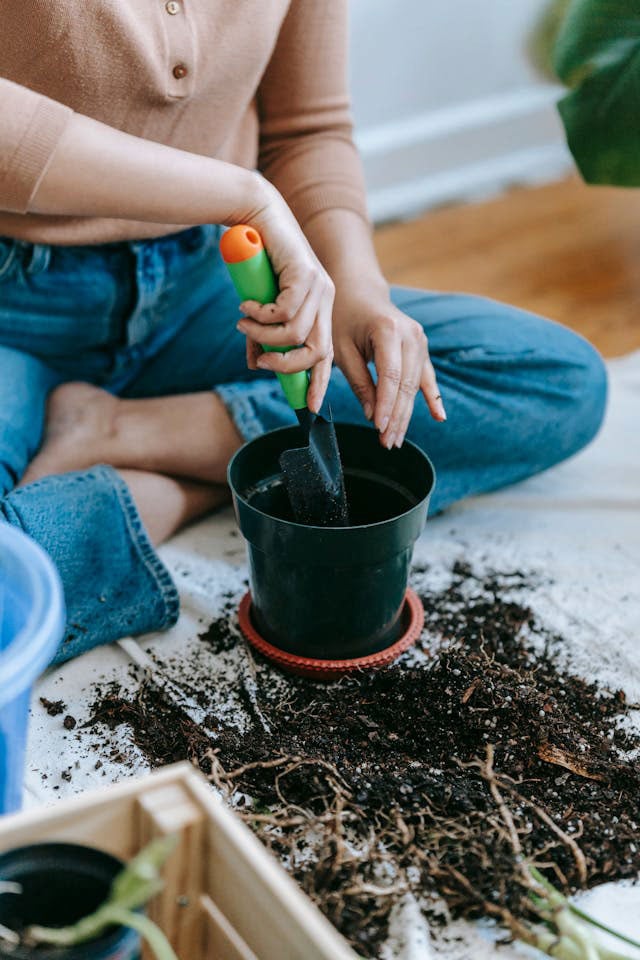 Close-up of a person sitting cross-legged holding a gardening trowel and digging into a small pot that is surrounded by soil and roots